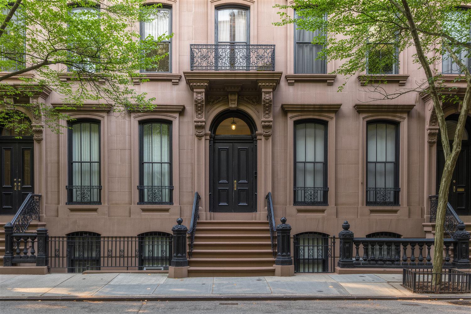 Brownstone row house facade with a stone stoop, iron railings and tall parlor windows beneath a street tree