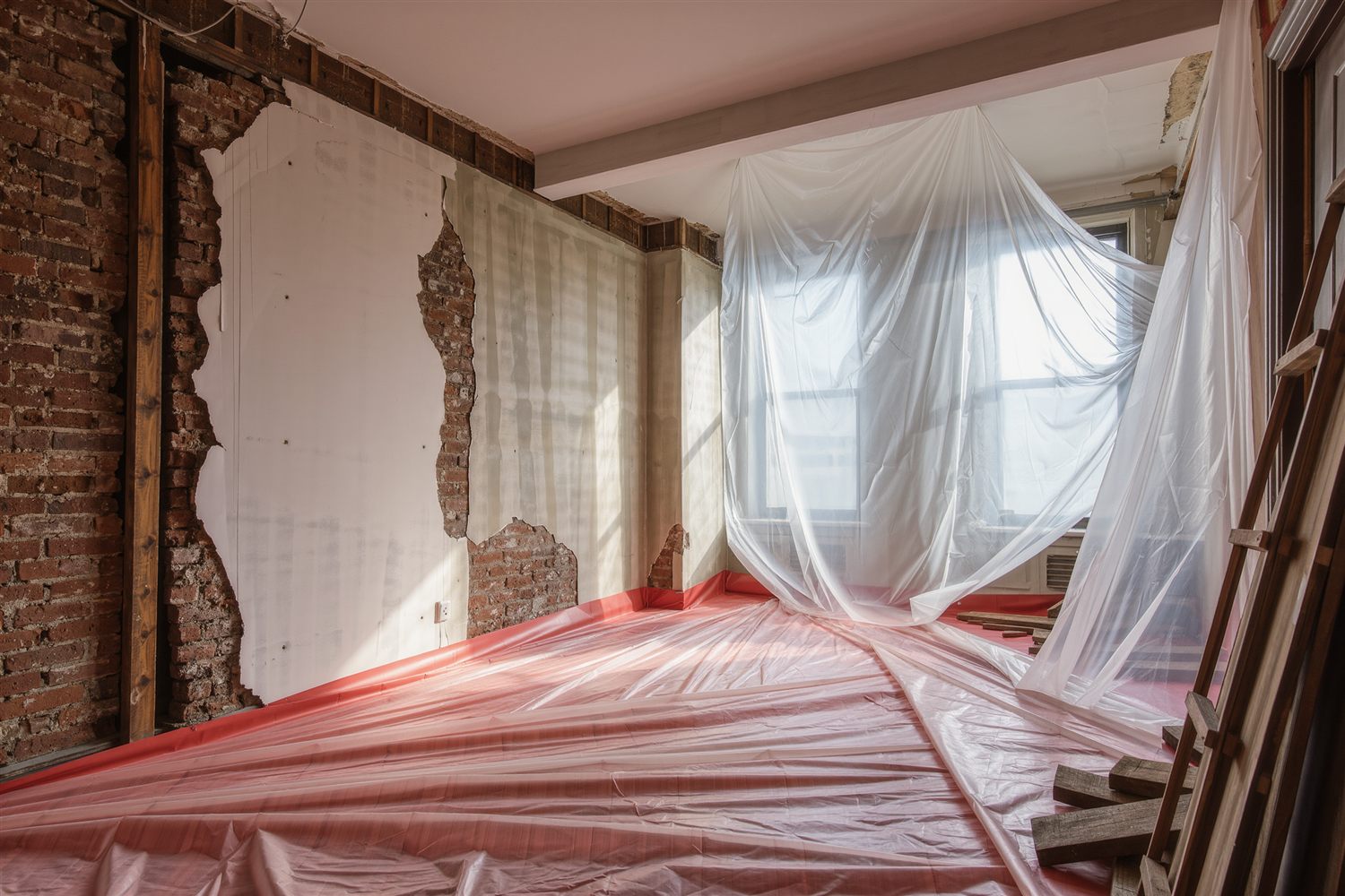 Gutted apartment room with plastic sheeting over the floor, an exposed brick wall and plaster stripped back