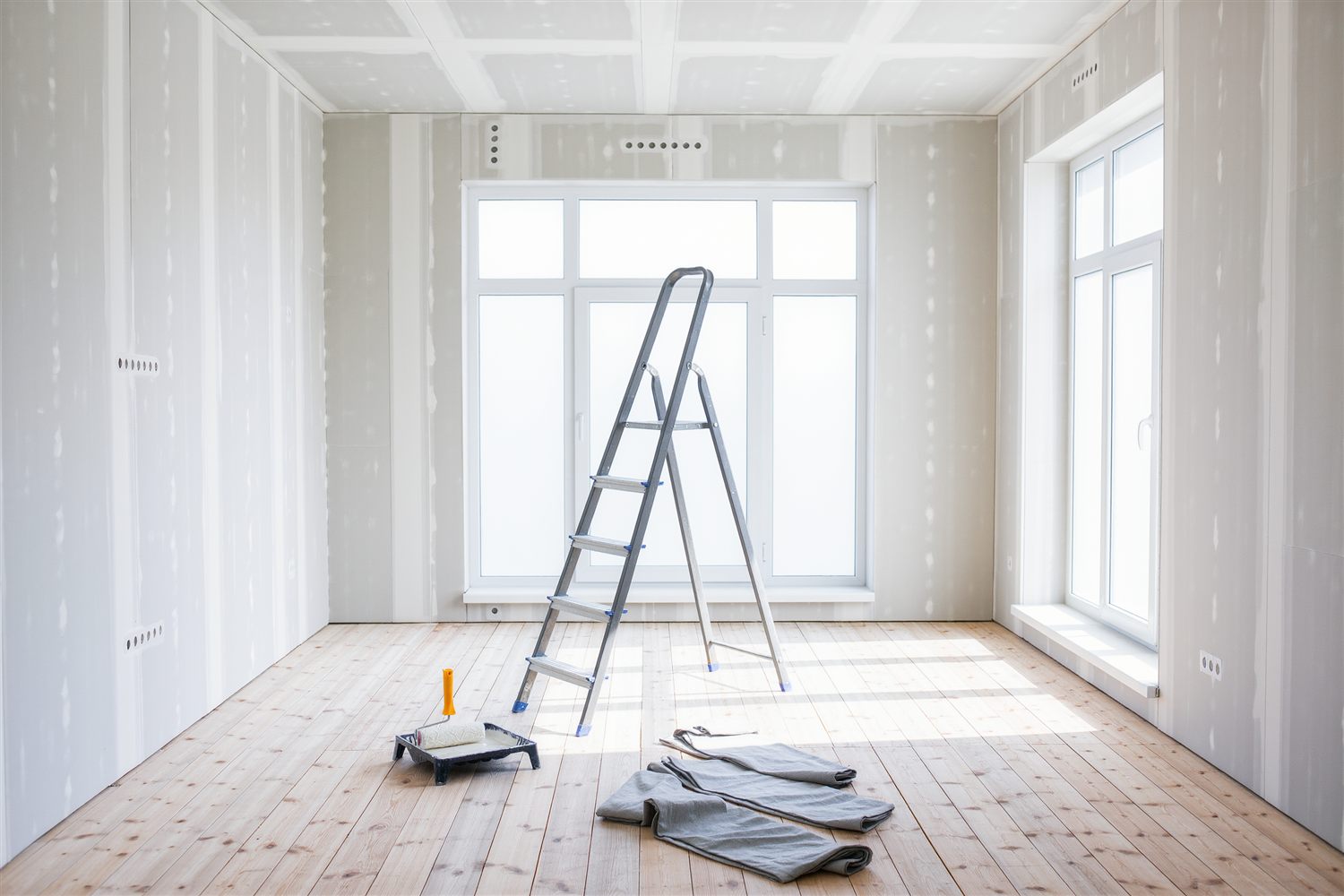 Empty room with taped drywall walls, a stepladder, a paint roller tray and drop cloths on a pine floor