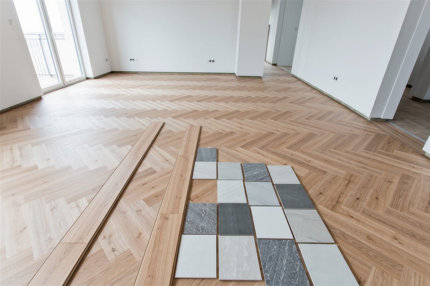 Herringbone wood planks being laid across an apartment floor with tile samples arranged beside them