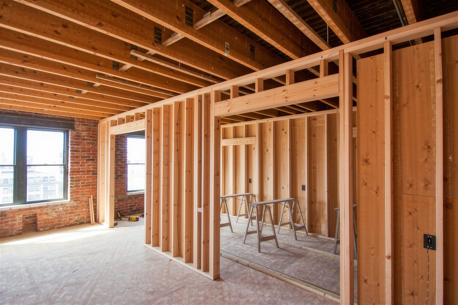 New wood stud framing inside an apartment with a bare brick wall, window light and sawhorses