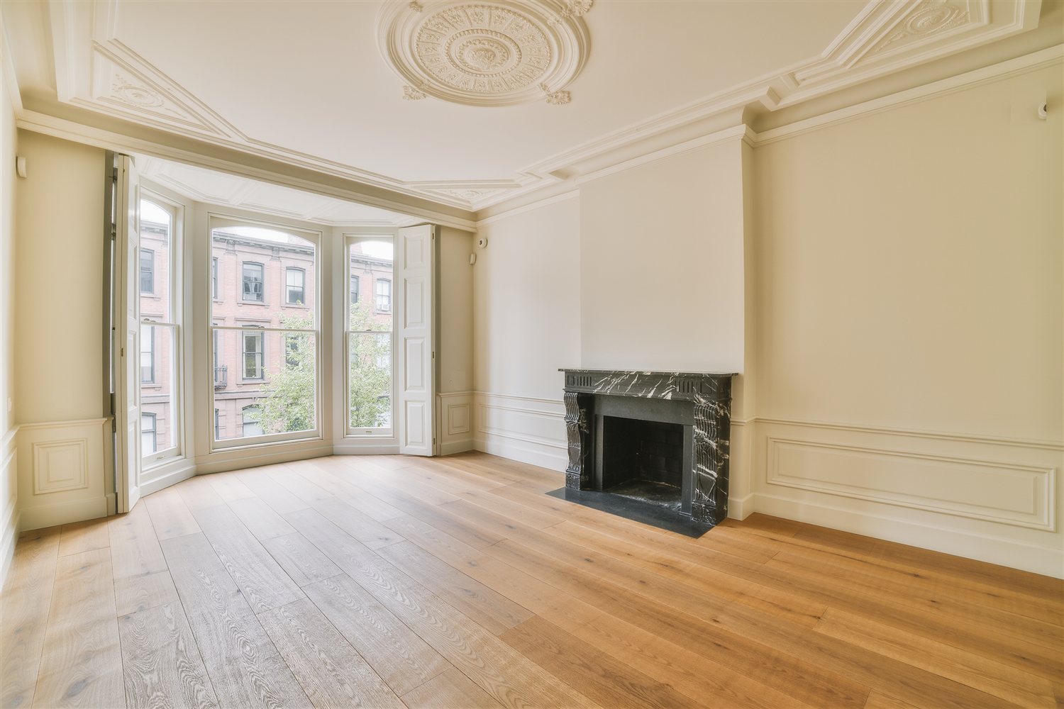 Empty parlor floor of a renovated New York brownstone with a bay window onto a brick-fronted street, an ornate plaster ceiling medallion, a black marble fireplace and wide oak floorboards